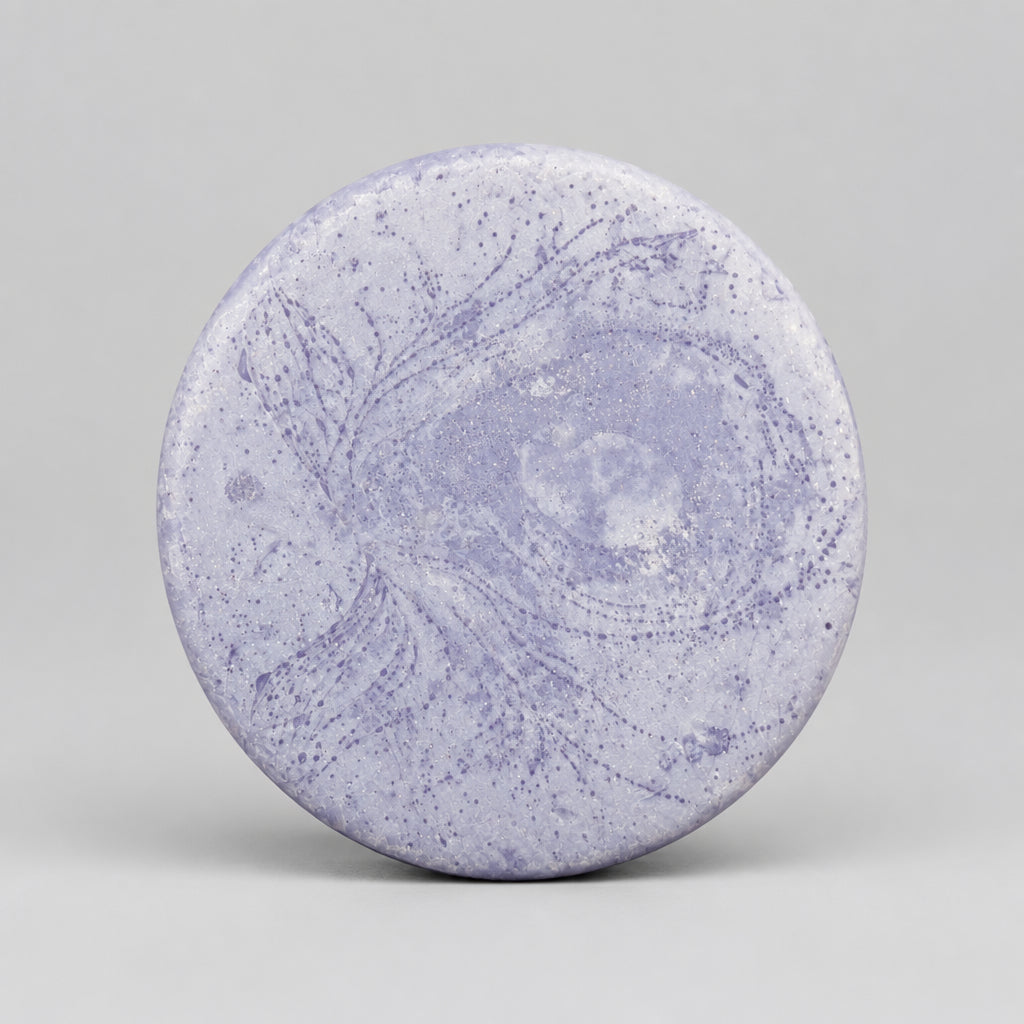 A round, light purple soap bar with a marbled swirl pattern, resting on a wooden surface.