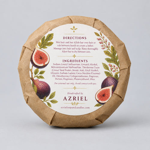 A round wrapped soap or bar product in brown kraft paper with a large white circular label featuring fig illustrations and the brand name Azriel. The label includes directions and ingredients text, photographed on a wooden surface.