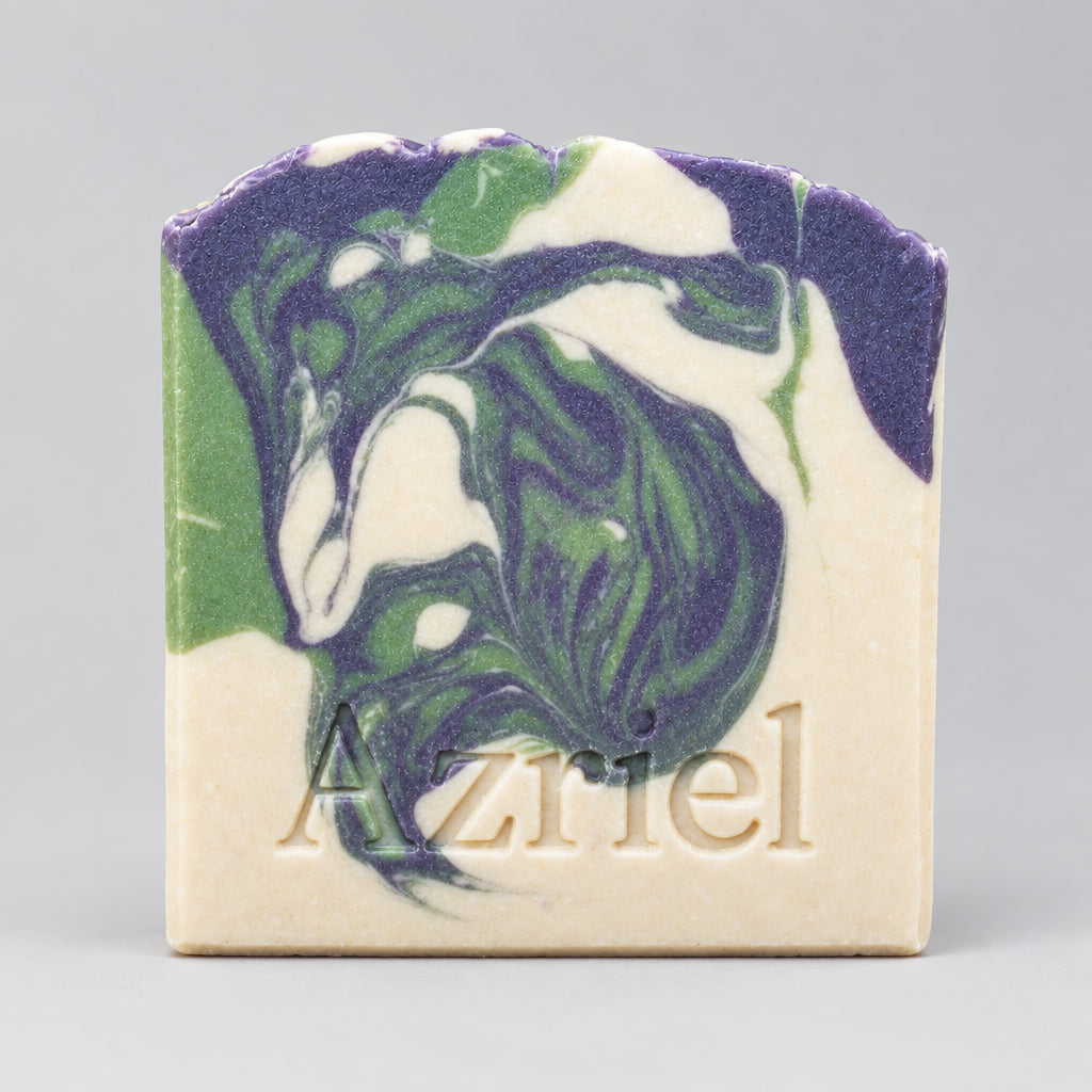 A handmade soap bar with a marbled swirl pattern of purple, green, and white on a cream base, featuring the name 'Azriel' embossed at the bottom, standing on a wooden surface against a dark background.