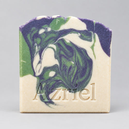 A handmade soap bar with a marbled swirl pattern of purple, green, and white on a cream base, featuring the name 'Azriel' embossed at the bottom, standing on a wooden surface against a dark background.